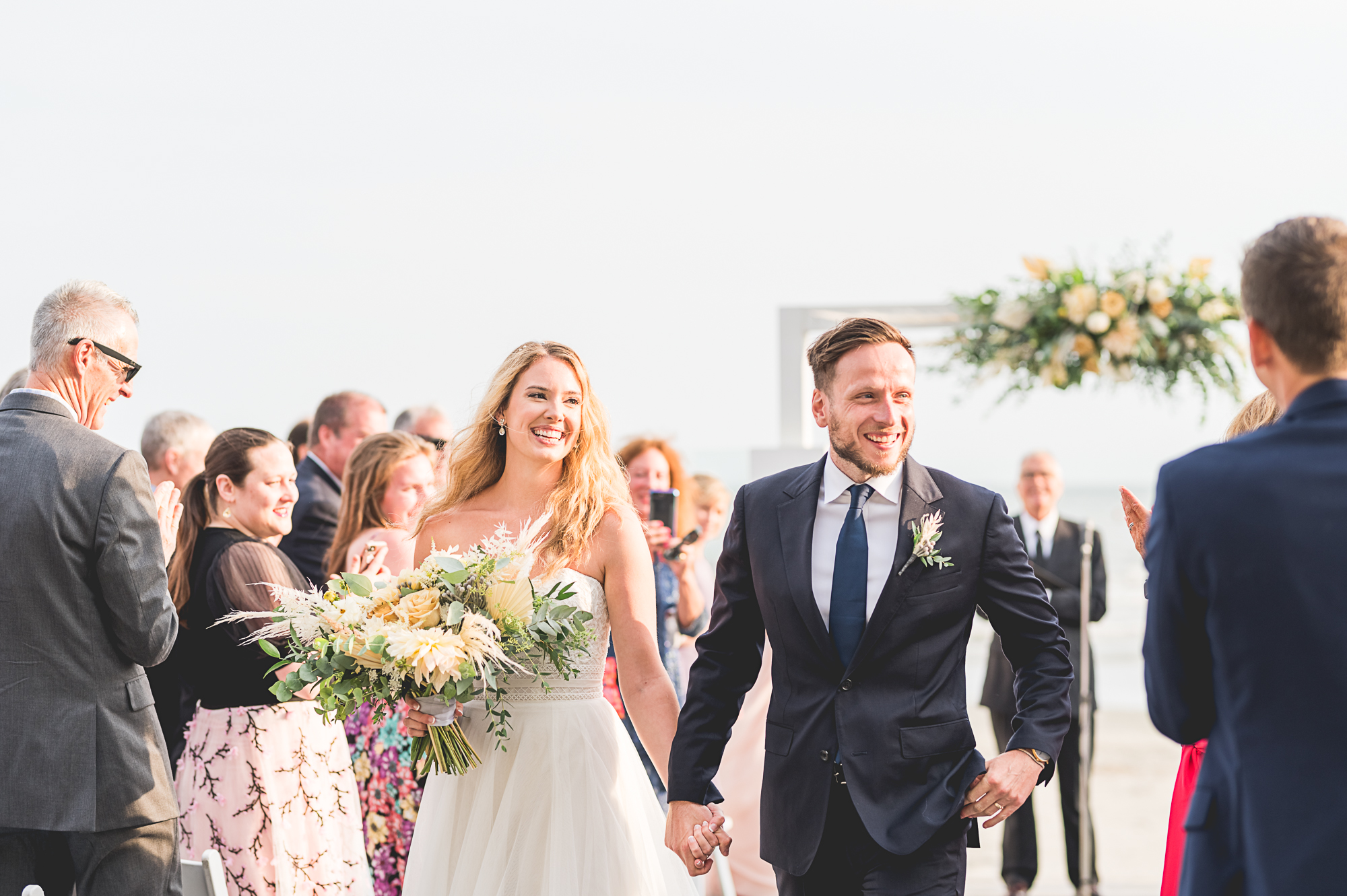 Wedding ceremony at Newport Beach House: A Longwood Venue in Newport, RI. Bride and groom smiling as they walk down the aisle holding hands.