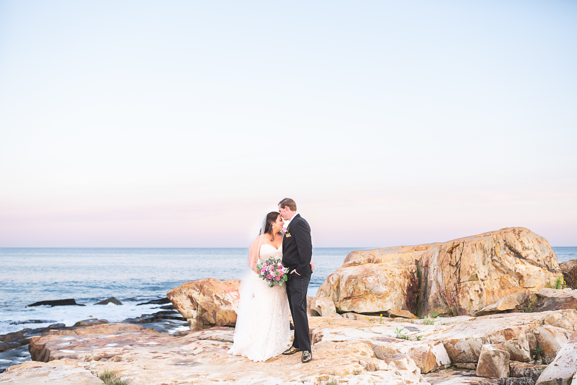 Wedding portraits in Bristol Rhode Island. Husband and wife standing on rocks by the ocean at sunset.
