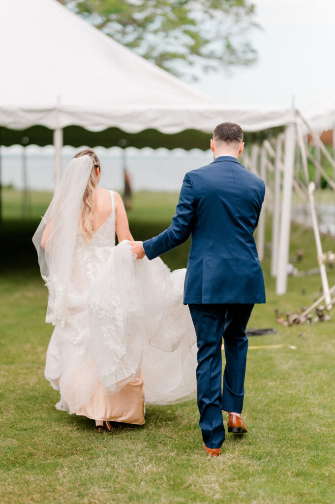A groom carrying his bride's dress walking across the grass to their reception tent at Marble House in Newport, Rhode Island.