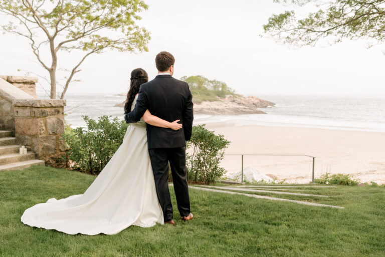 A bride and groom standing together, facing the ocean on their wedding day.